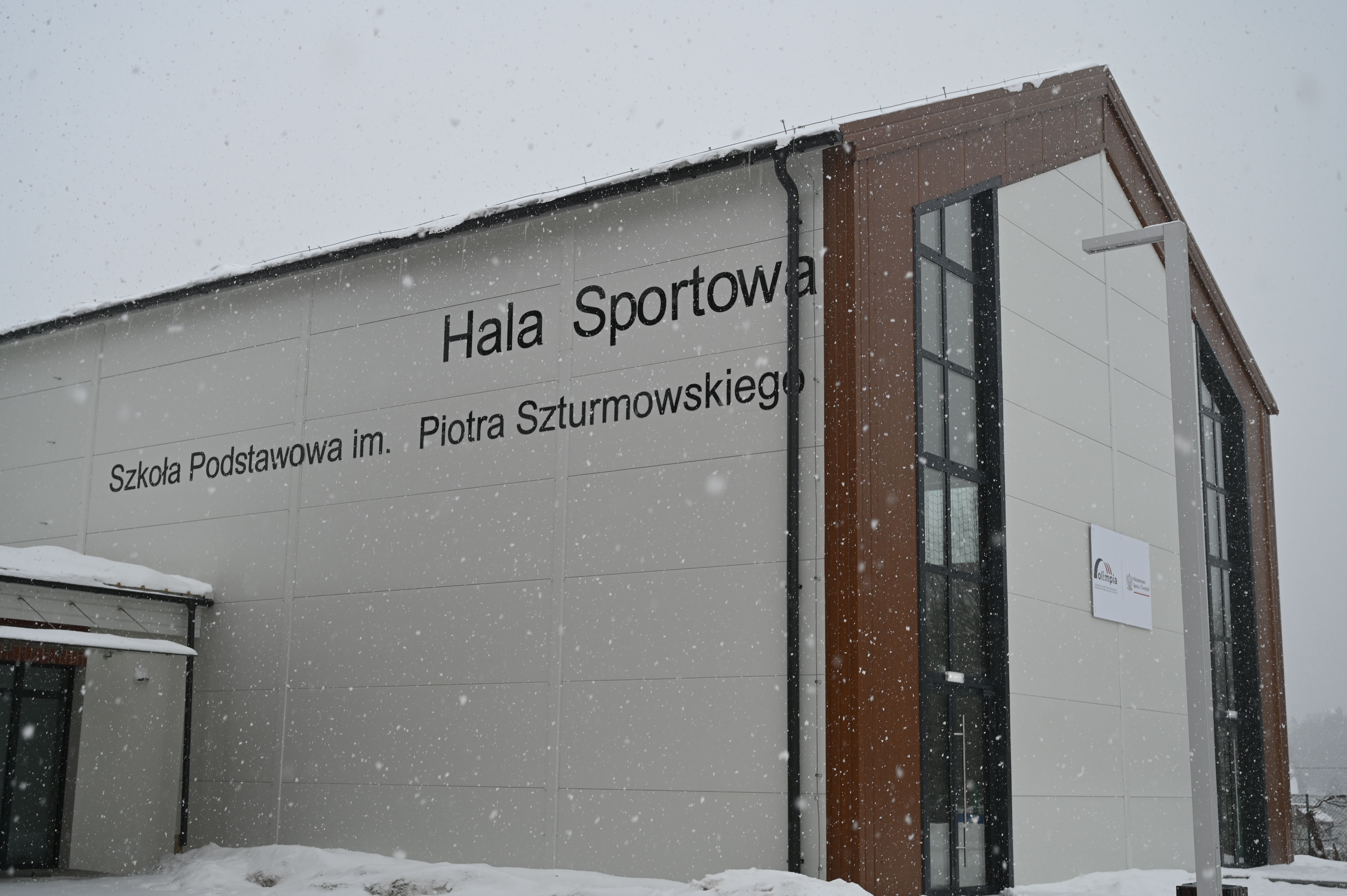 Nowoczesna hala sportowa szkoły podstawowej im. Piotra Szturmowskiego podczas opadów śniegu; jasna elewacja, duże okna i drewniane elementy wykończenia, wokół zalegający śnieg.